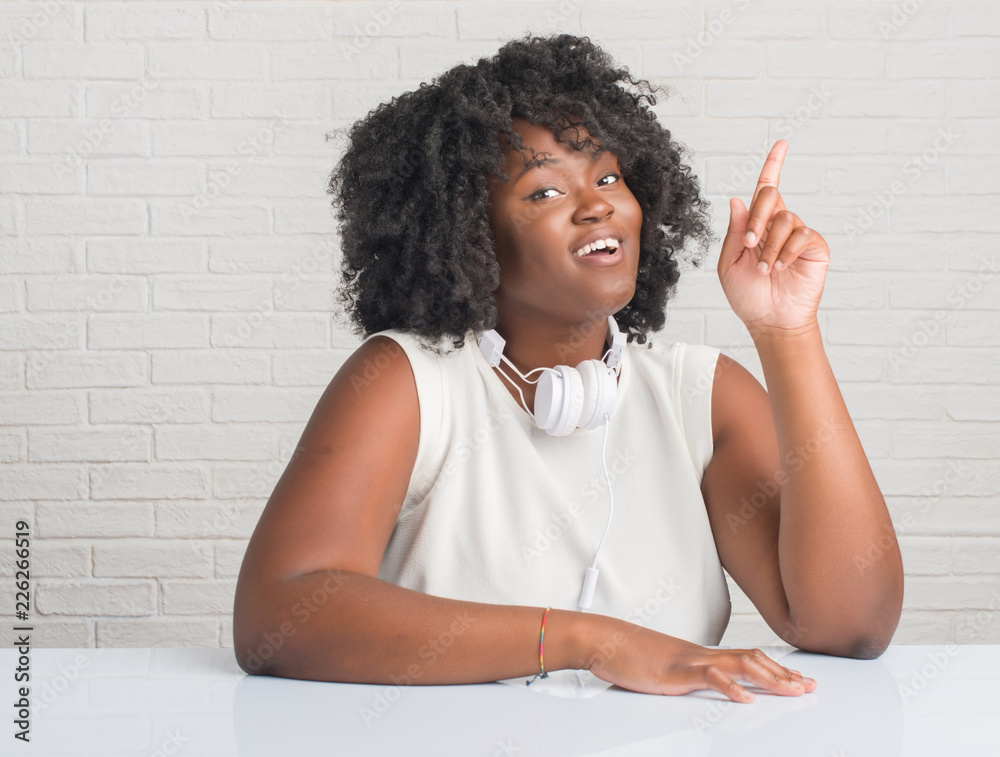 Young african american woman sitting on the table wearing headphones  pointing finger up with successful idea. Exited and happy. Number one.  Stock Photo | Adobe Stock