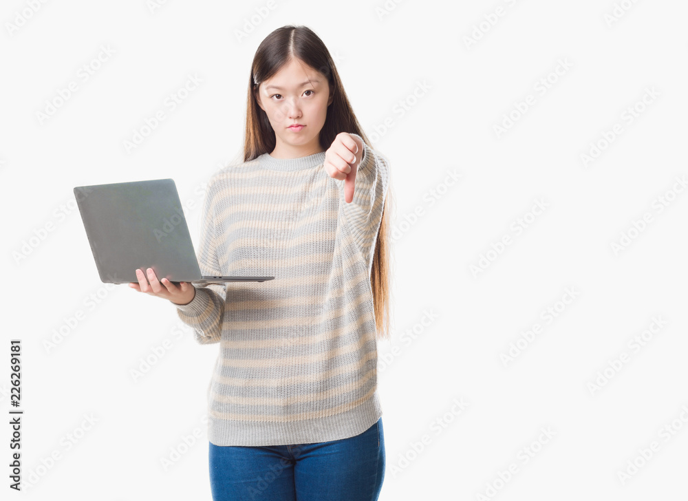Young Chinese woman over isolated background using computer laptop with angry face, negative sign showing dislike with thumbs down, rejection concept Young Chinese woman over isolated background using computer laptop with angry face, negative sign showing dislike with thumbs down, rejection concept