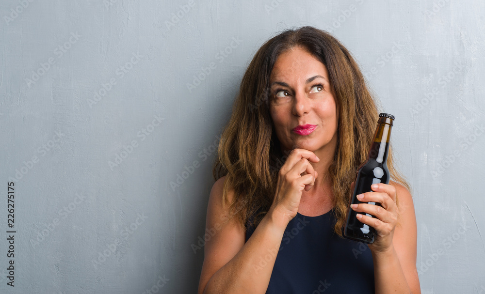Middle age hispanic woman standing over grey grunge wall holding beer bottle serious face thinking about question, very confused idea Middle age hispanic woman standing over grey grunge wall holding beer bottle serious face thinking about question, very confused idea