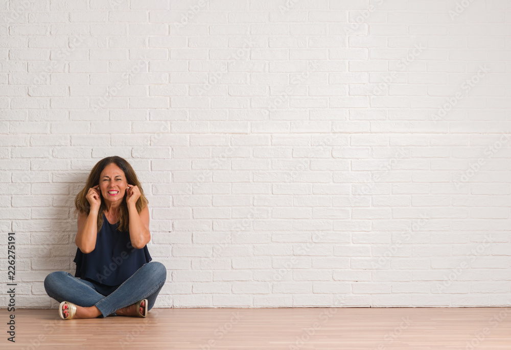 Middle age hispanic woman sitting on the floor over white brick wall covering ears with fingers with annoyed expression for the noise of loud music. Deaf concept. Middle age hispanic woman sitting on the floor over white brick wall covering ears with fingers with annoyed expression for the noise of loud music. Deaf concept.
