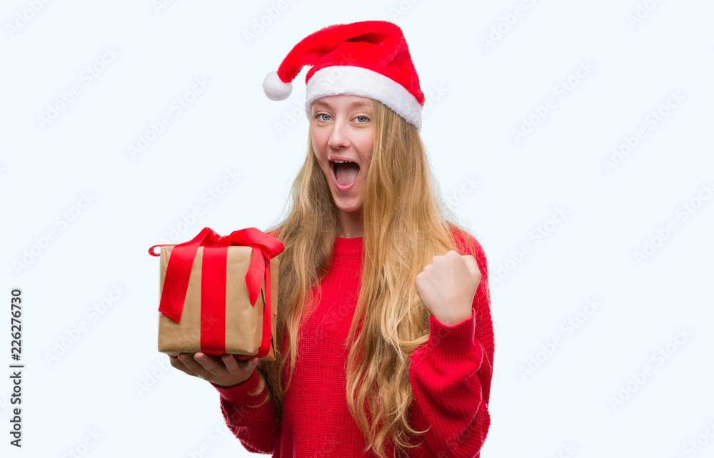 Blonde teenager woman wearing santa claus hat screaming proud and celebrating victory and success very excited, cheering emotion Blonde teenager woman wearing santa claus hat screaming proud and celebrating victory and success very excited, cheering emotion