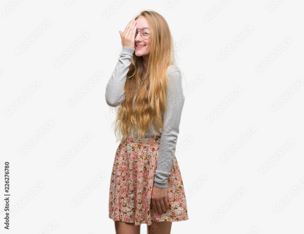 Blonde teenager woman wearing flowers skirt covering one eye with hand with confident smile on face and surprise emotion. Blonde teenager woman wearing flowers skirt covering one eye with hand with confident smile on face and surprise emotion.