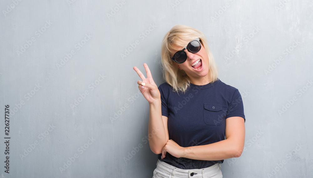 Adult caucasian woman over grunge grey wall wearing sunglasses smiling with happy face winking at the camera doing victory sign. Number two. Adult caucasian woman over grunge grey wall wearing sunglasses smiling with happy face winking at the camera doing victory sign. Number two.