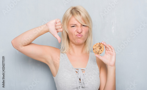 Caucasian adult woman over grey grunge wall eating chocolate cooky with angry face, negative sign showing dislike with thumbs down, rejection concept