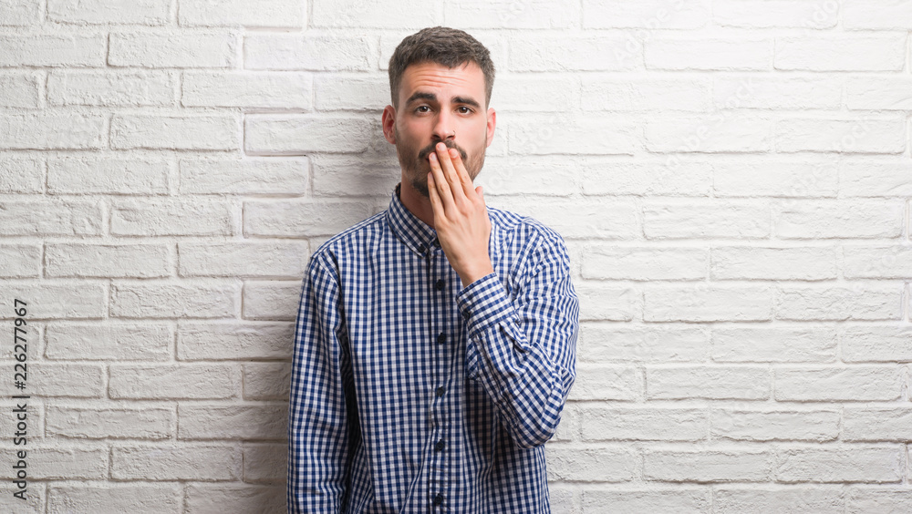 Young adult man standing over white brick wall cover mouth with hand shocked with shame for mistake, expression of fear, scared in silence, secret concept Young adult man standing over white brick wall cover mouth with hand shocked with shame for mistake, expression of fear, scared in silence, secret concept
