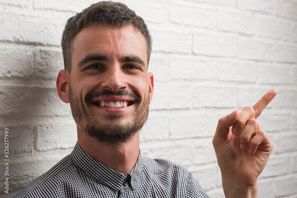 Young adult man standing over white brick wall very happy pointing with hand and finger to the side
