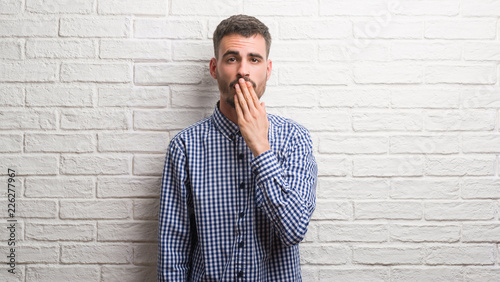 Young adult man standing over white brick wall cover mouth with hand shocked with shame for mistake, expression of fear, scared in silence, secret concept