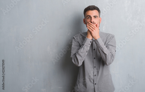Young adult man standing over grey grunge wall shocked covering mouth with hands for mistake. Secret concept.