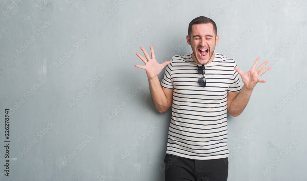 Young caucasian man over grey grunge wall celebrating mad and crazy for success with arms raised and closed eyes screaming excited. Winner concept Young caucasian man over grey grunge wall celebrating mad and crazy for success with arms raised and closed eyes screaming excited. Winner concept