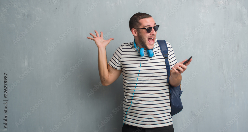 Young caucasian student man over grey grunge wall wearing headphones using smartphone very happy and excited, winner expression celebrating victory screaming with big smile and raised hands Young caucasian student man over grey grunge wall wearing headphones using smartphone very happy and excited, winner expression celebrating victory screaming with big smile and raised hands
