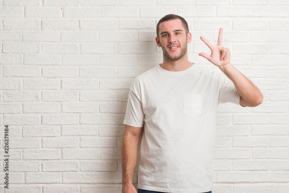 Young caucasian man standing over white brick wall showing and pointing up with fingers number three while smiling confident and happy. Young caucasian man standing over white brick wall showing and pointing up with fingers number three while smiling confident and happy.