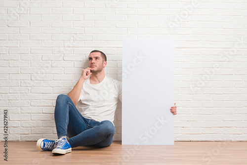 Young caucasian man sitting over white brick wall holding bottle of water serious face thinking about question, very confused idea