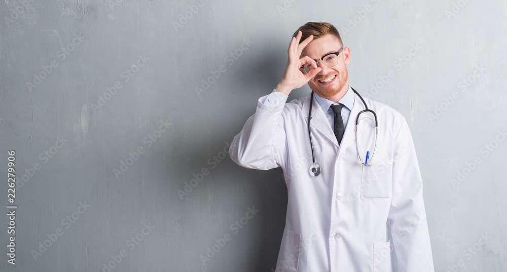 Young redhead doctor man over grey grunge wall with happy face smiling doing ok sign with hand on eye looking through fingers Young redhead doctor man over grey grunge wall with happy face smiling doing ok sign with hand on eye looking through fingers