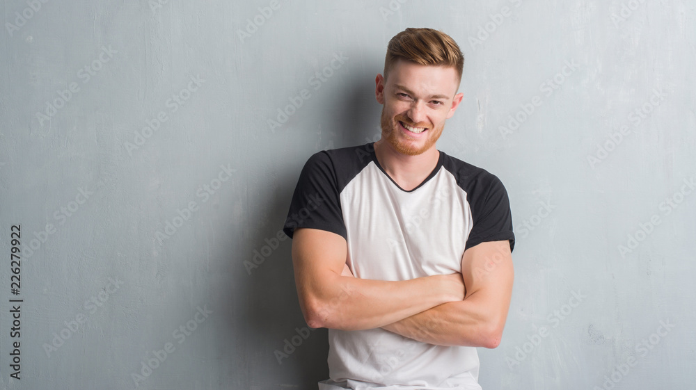 Young redhead man over grey grunge wall happy face smiling with crossed arms looking at the camera. Positive person.