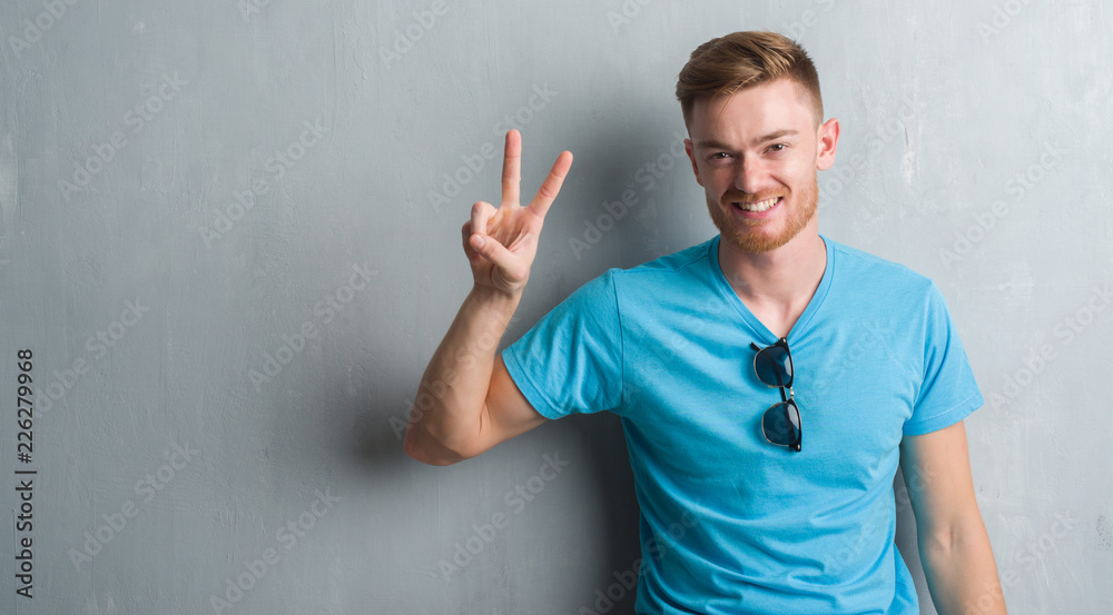 Fototapeta premium Young redhead man over grey grunge wall wearing casual outfit showing and pointing up with fingers number two while smiling confident and happy.