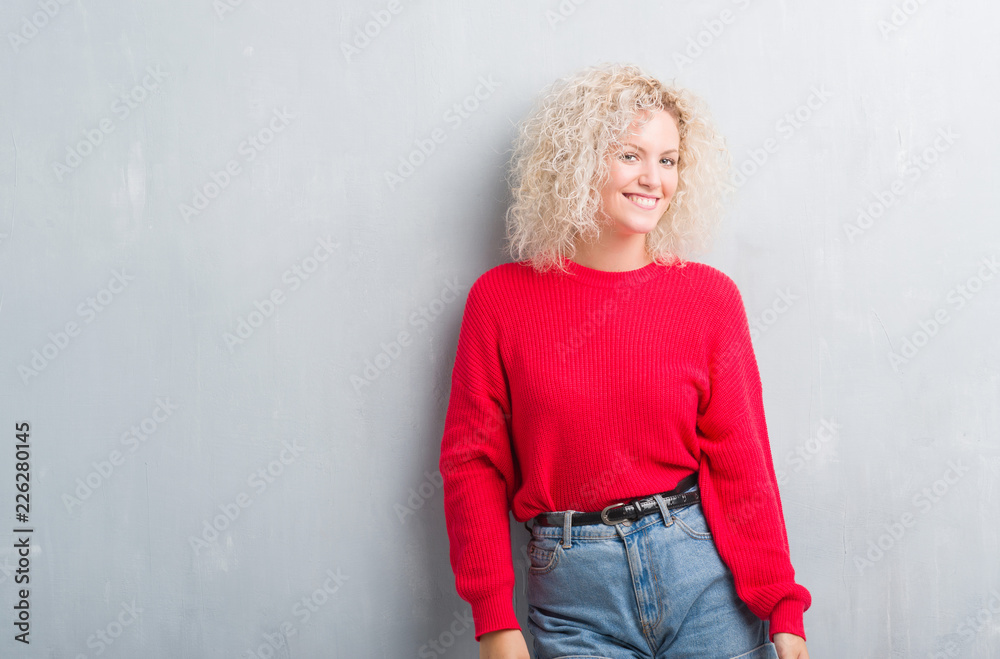 Young blonde woman with curly hair over grunge grey background looking away to side with smile on face, natural expression. Laughing confident. Young blonde woman with curly hair over grunge grey background looking away to side with smile on face, natural expression. Laughing confident.