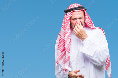 Senior arab man wearing keffiyeh over isolated background smelling something stinky and disgusting, intolerable smell, holding breath with fingers on nose. Bad smells concept. photo