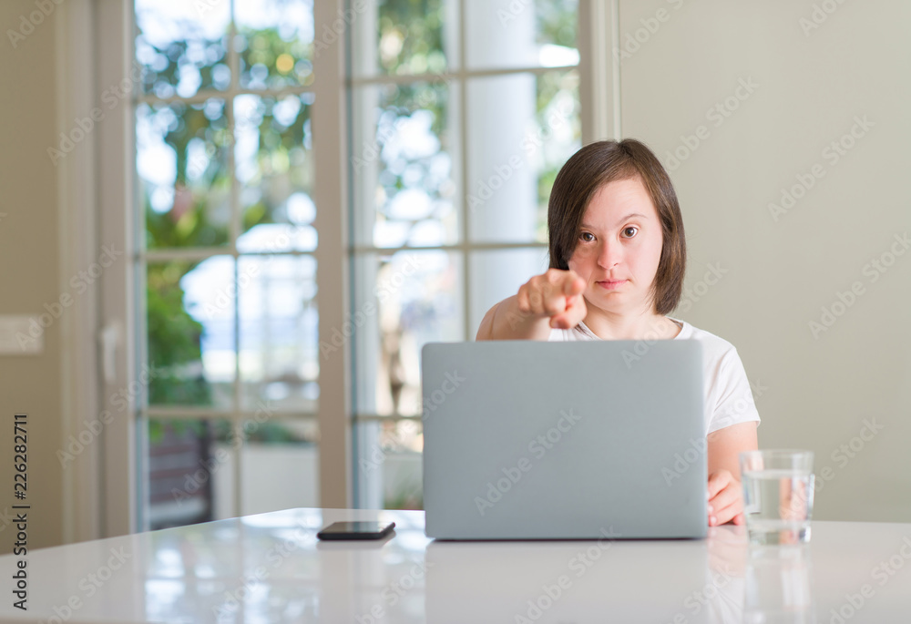 Down syndrome woman at home using computer laptop pointing with finger to the camera and to you, hand sign, positive and confident gesture from the front