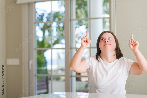 Down syndrome woman at home amazed and surprised looking up and pointing with fingers and raised arms.