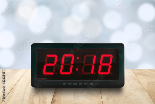 closeup red led light illuminated number 2018 on digital electric alarm clock face on brown wooden table top with defocused pastel blue and white Christmas lights bokeh background, time symbol concept