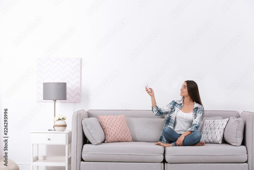 Young woman switching on air conditioner while sitting on sofa near white wall Young woman switching on air conditioner while sitting on sofa near white wall