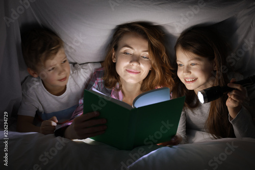 Mother reading bedtime story to her little children under blanket in evening