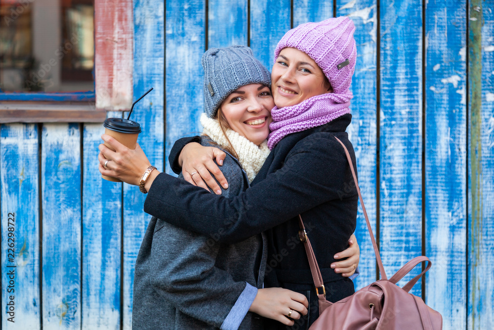 Obraz premium A young women friends in a stylish clothes hug, chat and drink coffee to go around blue wall. The concept of livestyle and outdoor recreation in autumn