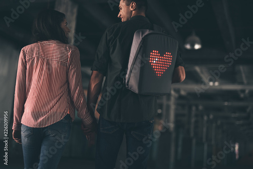 Heart image. Cheerful young man carrying an unusual backpack and smiling while looking at his beloved woman walking near
