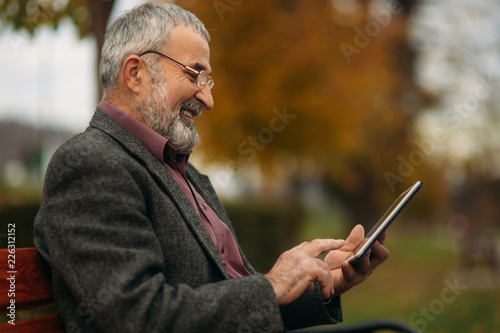 Grandpa uses a tablet and sits in the park on the bench. He look in to the screen and look photo