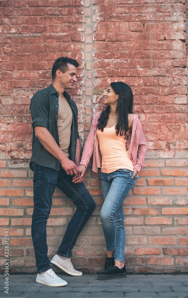 Brick wall. Romantic young man holding hand of his pretty smiling girlfriend and talking to her while standing next to the brick wall