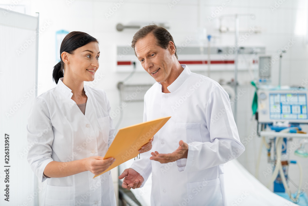 Waist up portrait of middle aged surgeon discussing patient illness history with colleague. Young lady looking at man with smile while holding documents Waist up portrait of middle aged surgeon discussing patient illness history with colleague. Young lady looking at man with smile while holding documents