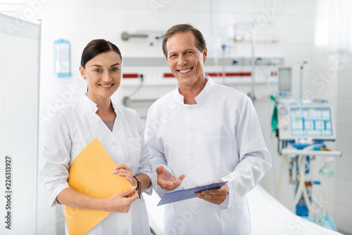 Waist up portrait of smiling health workers on blurred background. Lovely woman holding medical documents while confident man standing beside her and pointing hand at you