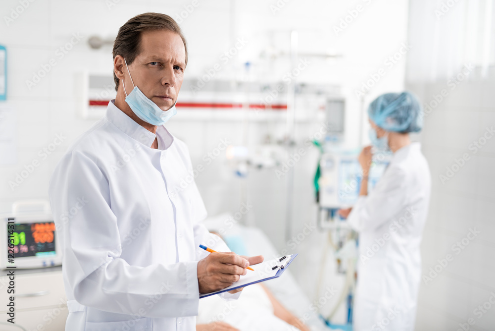 Waist up portrait of handsome surgeon with protective mask on chin noting information about patient while looking at camera with serious expression. Nurse near breathing machine on blurred background Waist up portrait of handsome surgeon with protective mask on chin noting information about patient while looking at camera with serious expression. Nurse near breathing machine on blurred background