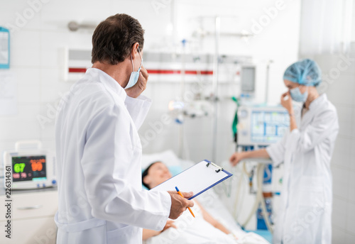 Back view portrait of surgeon in white lab coat holding clipboard and pen while touching protective mask on his face. Female medical worker talking with sick young lady on blurred background