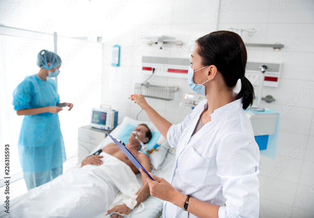 Lady in protective mask pointing at digital device for measuring heartbeat while nurse standing near patient. Middle aged gentleman on breathing machine sleeping in hospital bed