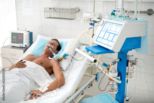 Portrait of unconscious middle aged man on mechanical ventilator lying in hospital bed under white sheets photo