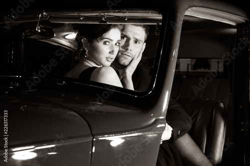 Good looking sexy couple, handsome man in suit, beautiful woman in red dress, are discovered embracing in vintage car