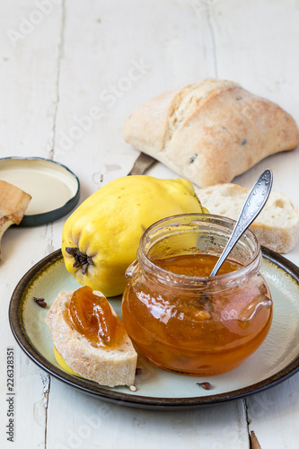A small glass jar of jam, a fresh ripe quince fruits and homemade white bread on an old table. 