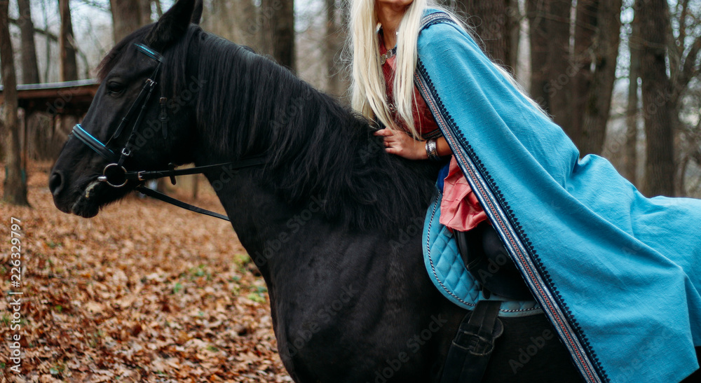 a girl with long hair, blonde hair, sits on the back of a black horse, in a blue cape and a vintage dress. throne, queen