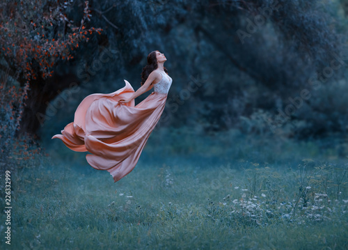 A young woman, a mysterious witch is floating in the air like a butterfly. A luxurious, long dress flutters in the wind in flight. Cold forest background. Enchanted princess. Art photo of levitation