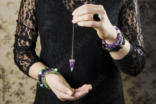 Close up of woman wearing black gothic clothing, hand holding and using amethyst crystal pendulum on silver chain fortune telling pendulum in hand. Mystical psychic dark witch concept.  photo