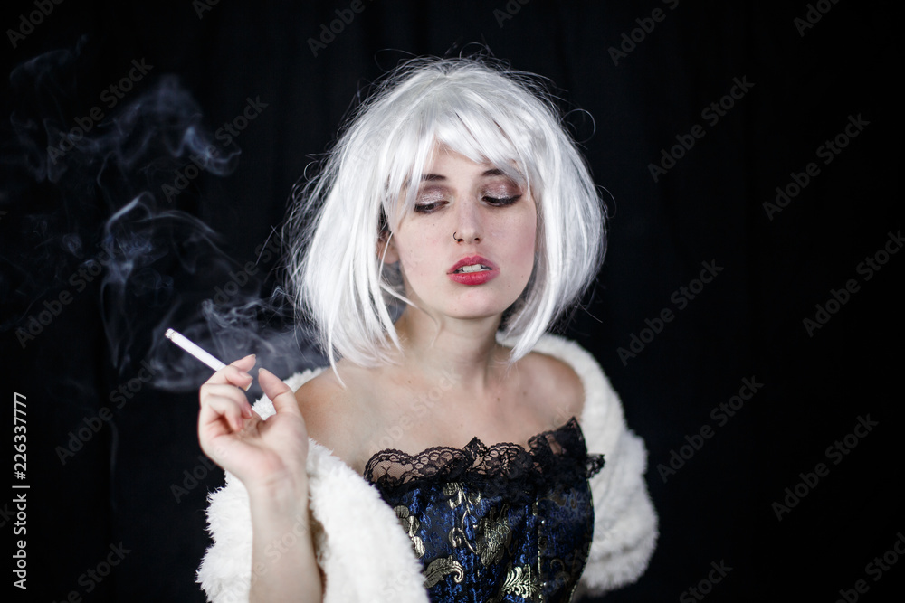 Attractive young woman in costume smoking cigar and looking at camera while standing on black background 