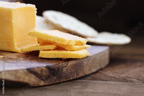 Block of cheddar cheese and slices over a rustic background.. Extreme shallow depth of field with selective focus on cheese. Water crackers in  blurred background.