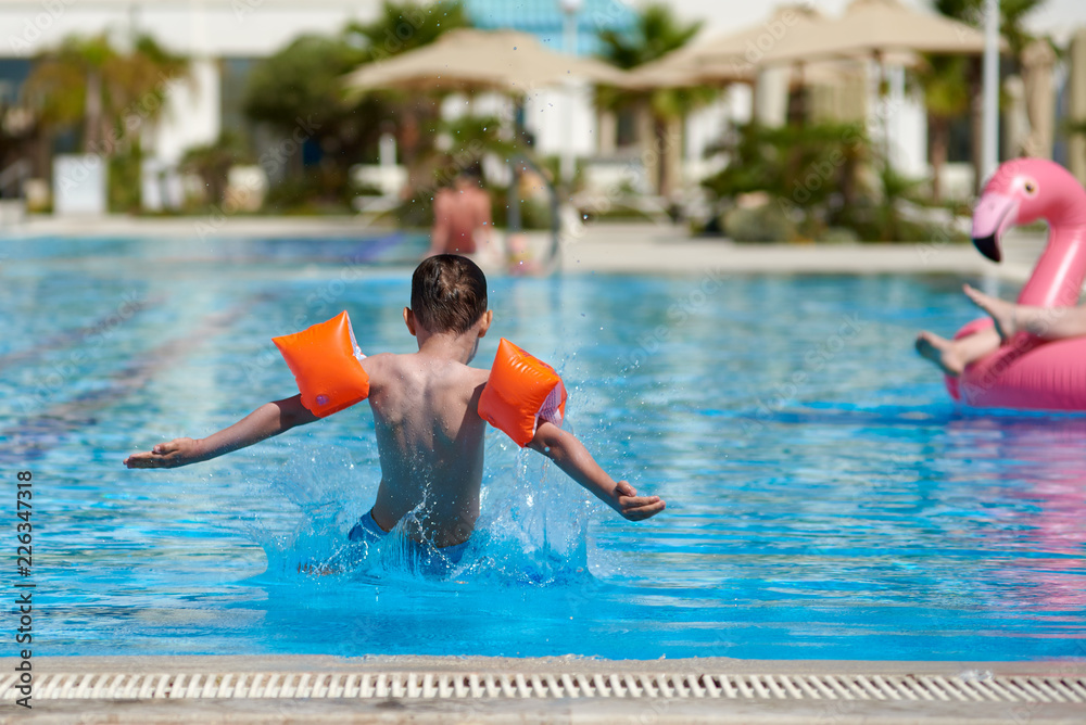 European boy in floating sleeves jumping into swimming pool at resort. He has wide open arms. Moment of entrance in water.