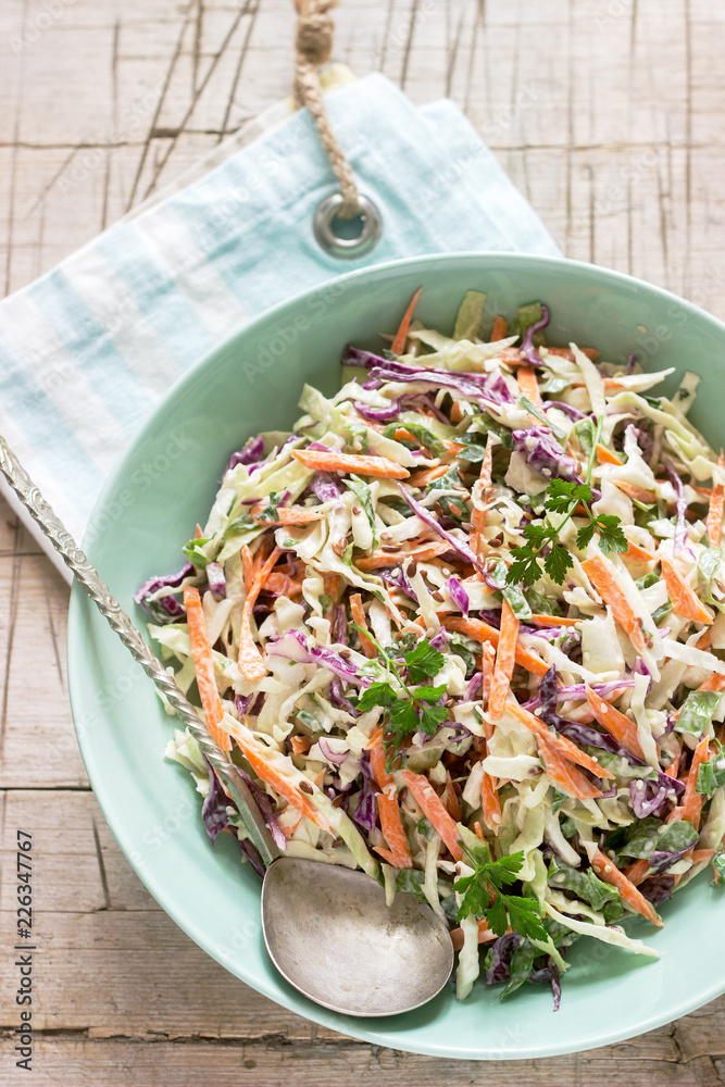 Coleslaw of cabbage, carrots and various herbs with mayonnaise in a large plate on a wooden background.
