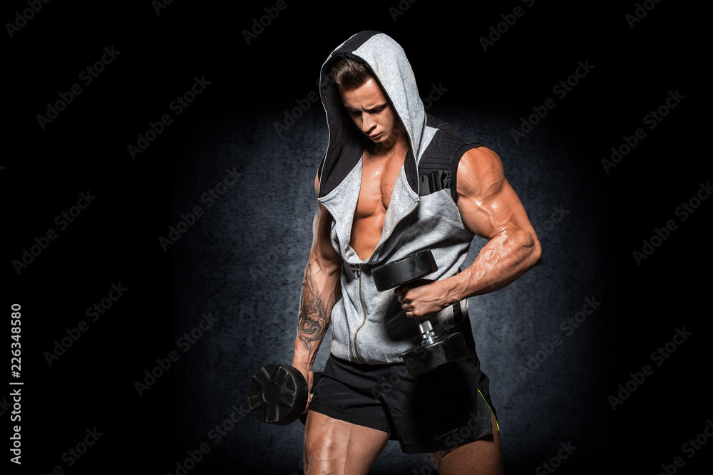 Young handsome athlete bodybuilder, in beautiful sportswear, demonstrates abdominals, against a dark background. Concept - power, beauty, biceps, triceps, sports equipment, gym, sports nutrition.