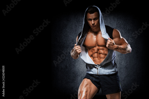 Young handsome athlete bodybuilder, in beautiful sportswear, demonstrates abdominals, against a dark background. Concept - power, beauty, biceps, triceps, sports equipment, gym, sports nutrition.