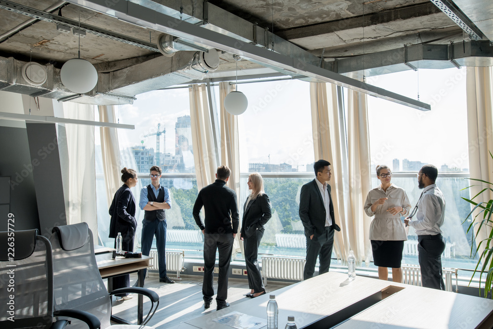 Group of modern business people standing at window with cityscape and discussing conference issues at break Group of modern business people standing at window with cityscape and discussing conference issues at break