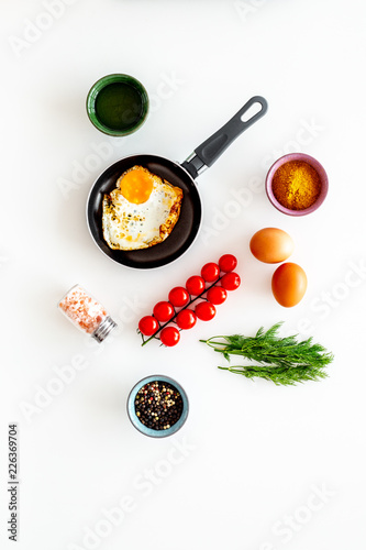 Cooking, preparing fried eggs with vegetables. Ready eggs in a frying pan near cherry tomatoes, greenery, spices, raw eggs on white background top view