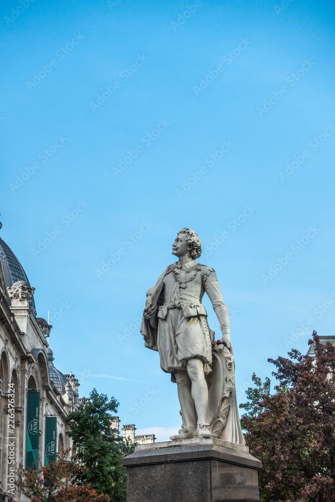 Antwerp, Belgium - September 24, 2018: Gray statue of Sir Anthony Van Dyck Flemish Painter against blue sky at Meir and Leysstraat link. Facades and some green foliage.